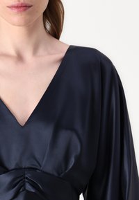 Tamsiai mėlyna atlasinė bluzonė su giliu V formos iškirpimu, surinktu perpus ties liemeniu ir ilgomis, laisvai krintančiomis rankovėmis, suteikiančiomis lygų paviršių.