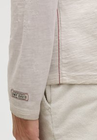 Langärmliges, hellbeiges Hemd aus strukturiertem Stoff. Mit rotem Akzentstich und einem "Camp David"-Logopatch auf dem Ärmel.