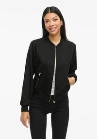 Junge Frau lächelnd, trägt eine schwarze Zip-Jacke mit goldenen Reißverschlüssen, ein weißes Hemd darunter und schwarze Hosen, steht vor einem weißen Hintergrund.