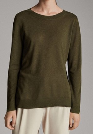 Kvinde iført en enkel olivengrøn langærmet trøje kombineret med lyse beige bukser foran en neutral baggrund.