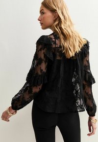 Svart spetsblus med volangärmar och blommönster. Genomskinligt tyg, avslappnad passform och eleganta detaljer. Modellen bär svarta byxor.