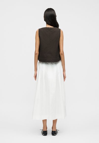 Καφέ αμάνικη μπλούζα με υφή, με φερμουάρ στην πλάτη, συνδυασμένη με μακρύ λευκό φόρεμα και μαύρα παπούτσια.