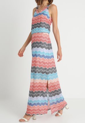 Femme portant une robe maxi sans manches à motifs avec des rayures en zigzag horizontales couleur corail, bleu, noir et blanc, associée à des sandales blanches à talons.