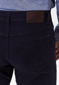 Pantaloni blu navy con una texture liscia, caratterizzati da una toppa in pelle marrone in vita e una tasca con dettagli di cucitura.