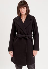 Manteau noir à ceinture avec col cranté, manches longues et poches latérales, fabriqué à partir d'un tissu doux et texturé, avec une coupe mi-longue.