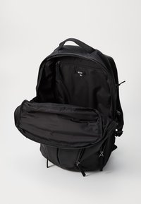 Sac à dos noir avec des compartiments zippés, fabriqué en tissu lisse. L'intérieur comprend un grand compartiment principal et une petite poche zippée.
