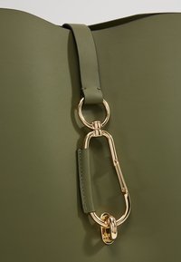 Olive zelená kožená kabelka se zlatým karabinovým zapínáním a kulatým kroužkem na uchu.