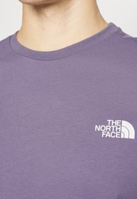 Krátký rukáv, tričko s kruhovým výstřihem v tlumené fialové barvě z bavlny s bílým logem na levém hrudníku. Hladká textura, klasický střih.