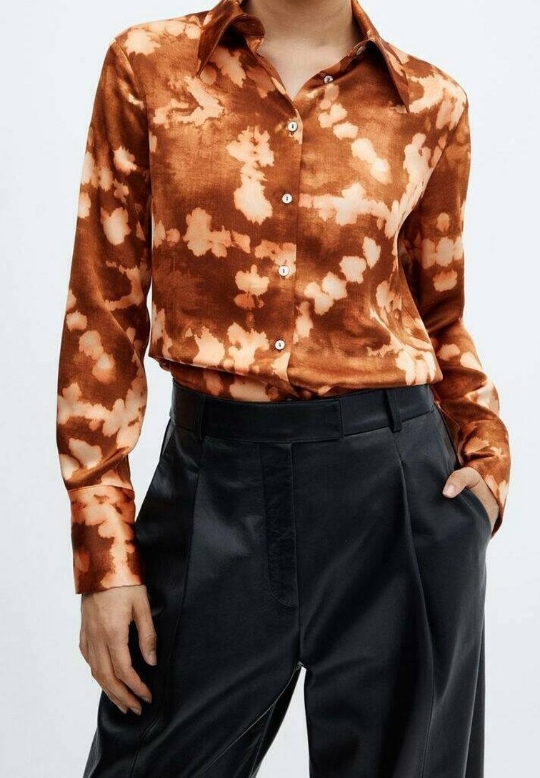 Donna che indossa una blusa a maniche lunghe con tie-dye ruggine e beige, infilata in pantaloni in pelle neri a vita alta, con una mano in tasca.