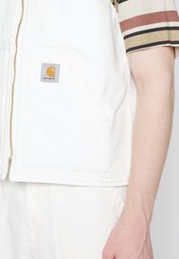 Weißer Canvas-Weste mit einem goldenen Reißverschluss und Tasche. Carhartt-Logo in Grau und Orange auf der linken Brust. Neutraler Hintergrund ohne Ablenkungen.