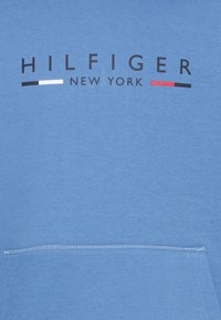 Šviesiai mėlynas džemperis su kengūros kišene. Puošia juodas ir raudonas „HILFIGER NEW YORK“ logotipas. Pagamintas iš minkštos medžiagos su lygia tekstūra.