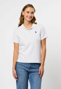 Hvit polo-skjorte med ribbet tekstur, korte ermer og knappestolpe med tre knapper. Har en liten marineblå logo på venstre bryst.