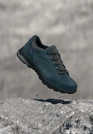 Blauer Wanderschuh mit schwarzer Sohle und Schnürsenkeln, der über felsiges Terrain schwebt und kleine Trümmer vor einem verschwommenen grauen Hintergrund aufwirbelt.