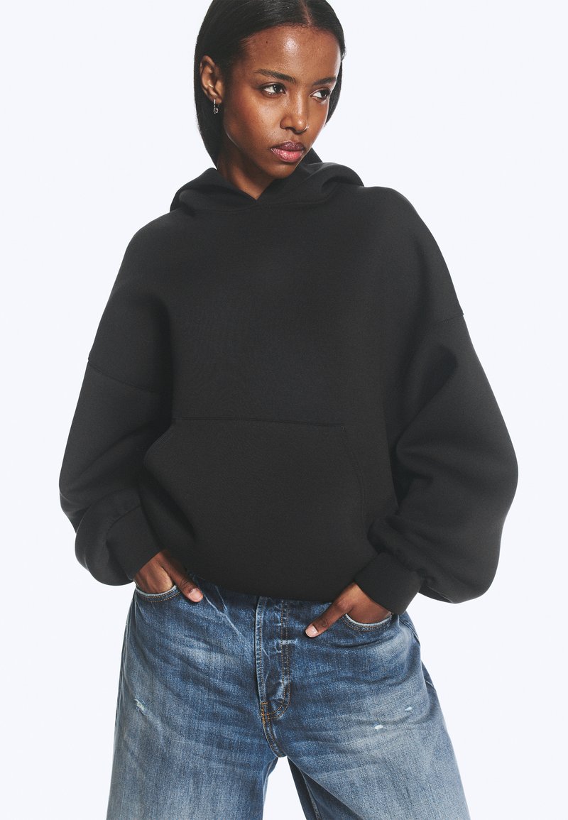 Kvinde iført en sort oversized hættetrøje med hænder i lommerne og løstsiddende blå jeans, kigger til siden mod en ensfarvet baggrund.