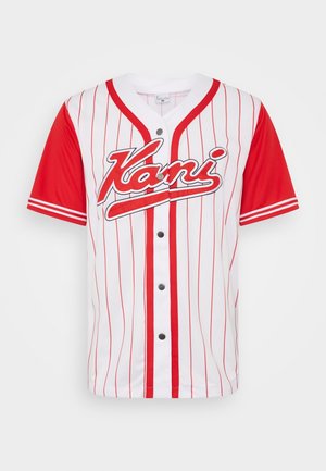 Person trägt ein weiß-rotes Vans-Baseball-Trikot mit vertikalen Streifen und mehrfarbigen gestreiften Hosen, steht neben einer Metallstruktur.