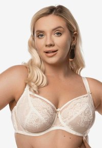 Koronkowy biustonosz w kolorze kremowym z fiszbinami, przezroczystymi panelami i delikatnym detalem w postaci kokardki na środku. Regulowane ramiączka. Gładka tekstura.