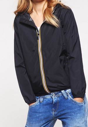 Woman wearing a black lightweight hooded jacket with striped zipper and blue jeans, hands in pockets, against a plain background.