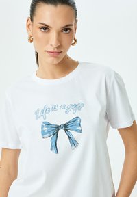Weißes Baumwoll-T-Shirt mit blauem grafischen Text "Das Leben ist ein Geschenk" und einer Bow-Illustration. Kurze Ärmel und Rundhalsausschnitt.