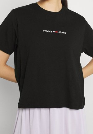Persoon draagt een zwart Tommy Jeans t-shirt met een wit en rood logo gecentreerd op de borst, gecombineerd met een lichtgekleurde rok of broek.