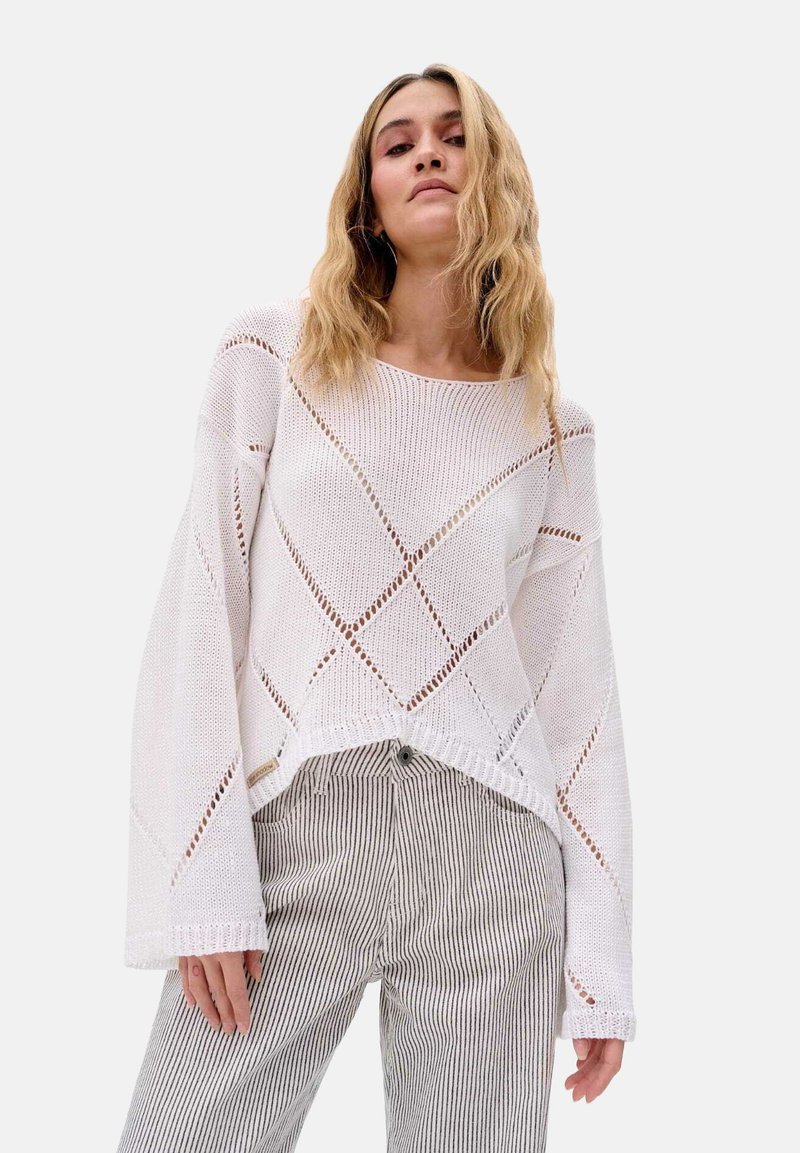 Weißer, strukturierter Strickpullover mit lockerer Passform, der über diamantförmige Muster und weite Ärmel verfügt. Kombiniert mit gestreiften, hochgeschnittenen Hosen.