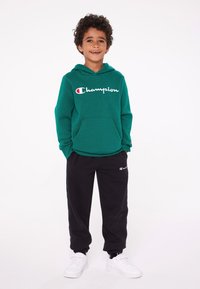 Felpa verde con logo "Champion", tasca frontale, pantaloni sweat neri con logo, sneakers bianche; tutti su uno sfondo neutro.