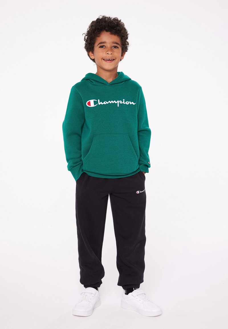 Felpa verde con logo "Champion", tasca frontale, pantaloni sweat neri con logo, sneakers bianche; tutti su uno sfondo neutro.