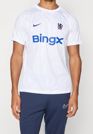 Man wearing white Nike Chelsea FC soccer jersey with blue Bingx logo and navy blue Nike pants, standing against plain background.