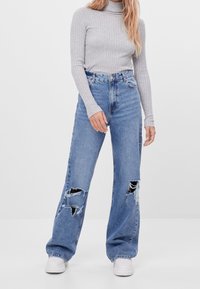 Femme portant un pull à col roulé côtelé gris, un jean large taille haute déchiré bleu et des baskets blanches, se tenant devant un fond uni.