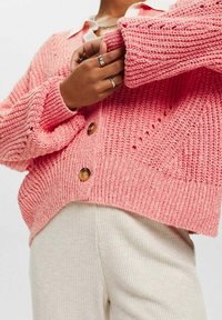 Cardigan en tricot rose avec une coupe décontractée, dotée de fermetures à bouton et de poignets côtelés, associé à un pantalon crème côtelé.