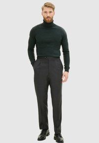 Grüner Rollkragenpullover kombiniert mit grauen maßgeschneiderten Hosen. Die Hose hat einen einzelner Knopfverschluss und seitliche Taschen. Schwarze Schuhe runden das Outfit ab.