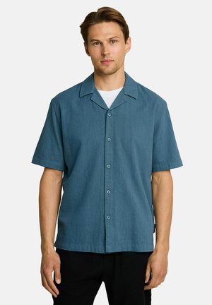 Man wearing a blue short-sleeve button-up shirt over a white t-shirt, standing against a plain light background.