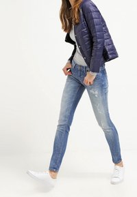 Blue distressed skinny jeans with faded areas, paired with a gray top and a navy quilted jacket. Footwear is white sneakers.
