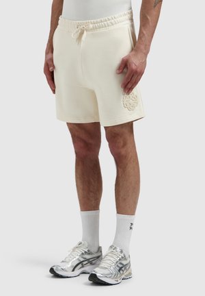 Männliches Model trägt cremefarbene Shorts mit Kordelzug und geprägtem Logo am linken Bein, weiße Sportsocken und silberne Laufschuhe, steht vor grauem Hintergrund.