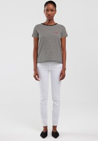 Camiseta a rayas en blanco y negro, de manga corta, con cuello redondo, que presenta un pequeño logo rojo. Combinada con pantalones ajustados blancos y zapatillas negras sin cordones.
