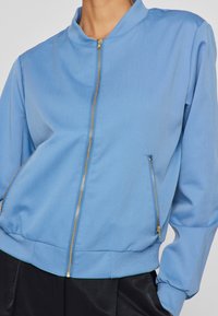 Svjetloplavi bomber jakna s patentnim zatvaračem, rebrastim ovratnikom i manžetama. Sadrži džepove s patentnim zatvaračem i struk prilagođen tijelu. Glatka tekstura tkanine.