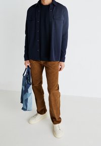 Laivastonsininen paidantakki, jossa on kaksi rintataskua, kerroksittain mustan t-paidan päällä, yhdistetty ruskeisiin samettihousuihin ja valkoisiin lenkkareihin. Mukana sininen laukku.