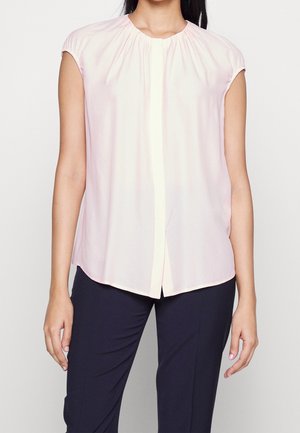 Femme portant un chemisier sans manches rose clair avec un col froncé et un pantalon droit foncé, debout devant un fond uni.