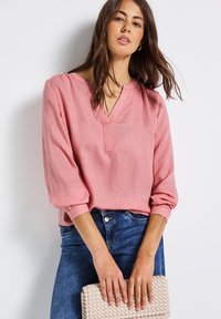 Blusa lunga manica rosa chiaro con scollatura a V, polsini elasticizzati e tessuto strutturato. Abbinata a jeans blu e una pochette intrecciata beige.