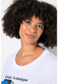 Camiseta de algodón blanca con escote redondeado que presenta un texto gráfico en negro y azul. La modelo tiene el cabello corto y rizado de color oscuro y un maquillaje natural.