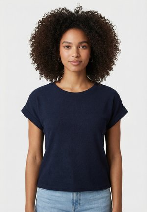 Femme aux cheveux bouclés et foncés, portant un haut bleu marine à manches courtes et un jean bleu clair, debout devant un fond uni clair.