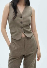 Brązowa bezrękawnik z dekoltem w kształcie V, wyposażona w pięć guzików i kieszenie z przodu, w połączeniu z dopasowanymi spodniami w podobnym materiale i fakturze.