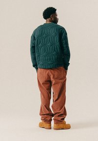 Dunkelgrüner, gestepptes Sweatshirt mit einem strukturierten Muster, kombiniert mit lockeren, karierten rustfarbenen Hosen und hellbraunen Schnürstiefeln.