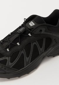 Fekete sportcipő, amely hálós és szintetikus anyagokból készült, texturált felülettel, ívelt kialakítással, visszatükröződő díszítésekkel és rugalmas fűzőkkel.