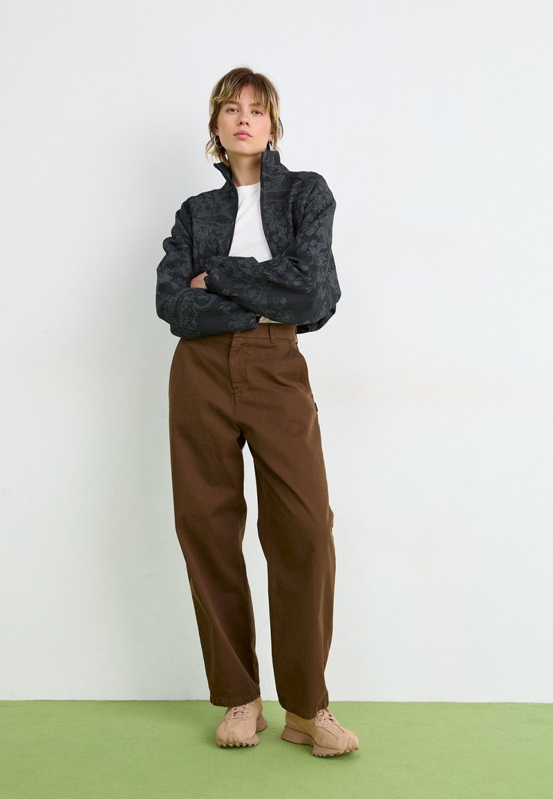 Veste noire à motifs avec col haut, chemise blanche en dessous, pantalon marron et chaussures marron clair, se tenant sur un sol vert.