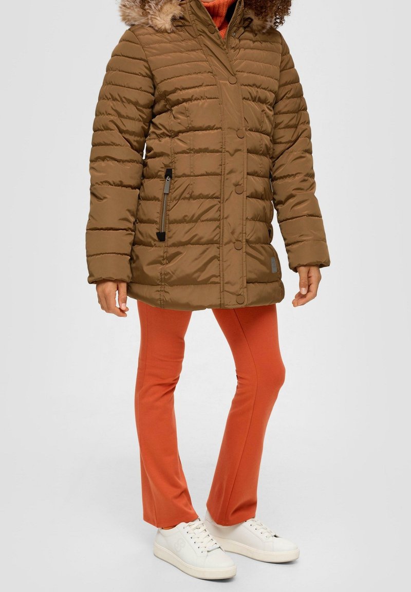 Femme portant une doudoune marron avec une capuche bordée de fourrure, pantalon orange et baskets blanches, debout devant un fond uni.