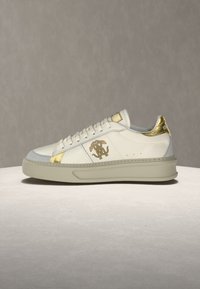 Λευκό χαμηλό αθλητικό παπούτσι με μπεζ σόλα, χρυσές λεπτομέρειες στη φτέρνα, στην περιοχή των κορδονιών και στο λογότυπο στο πλάι, τοποθετημένο σε επίπεδη επιφάνεια προς απλό φόντο.