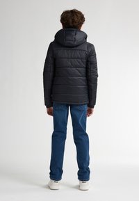 Zwarte pufferjack met een capuchon, voorzien van gestikte panelen en elastische manchetten, gecombineerd met blauwe jeans en witte sneakers.