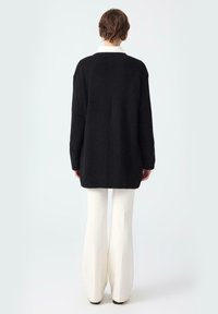 Person standing facing away wearing a long black ribbed sweater and white wide-leg pants against a plain background.