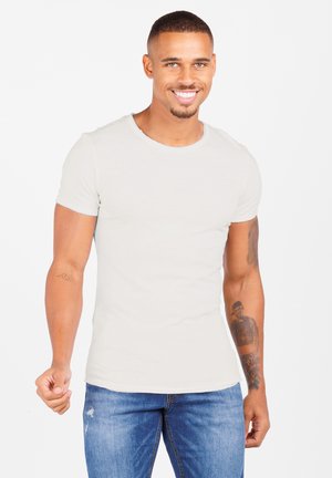Smiling man wearing a light grey t-shirt and blue jeans, standing against a plain white background and making a fist with his right hand.