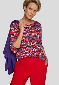 Bunter gemusterter Oberteil in Rot, Lila und Schwarz, mit Rundhalsausschnitt und Schleifendetail. Modell trägt rote Hosen und hält einen lila Pullover.